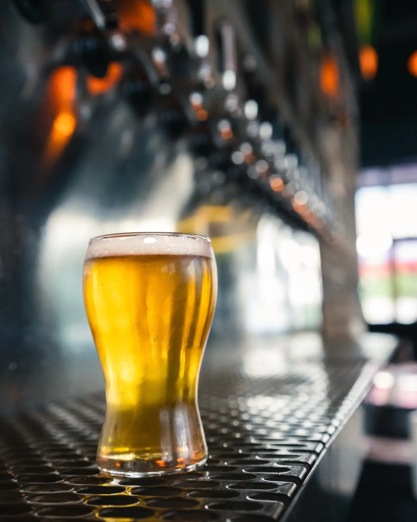 A glass of golden draft beer on the bar mat beneath Celebration Brewing Co.'s chrome tap line.