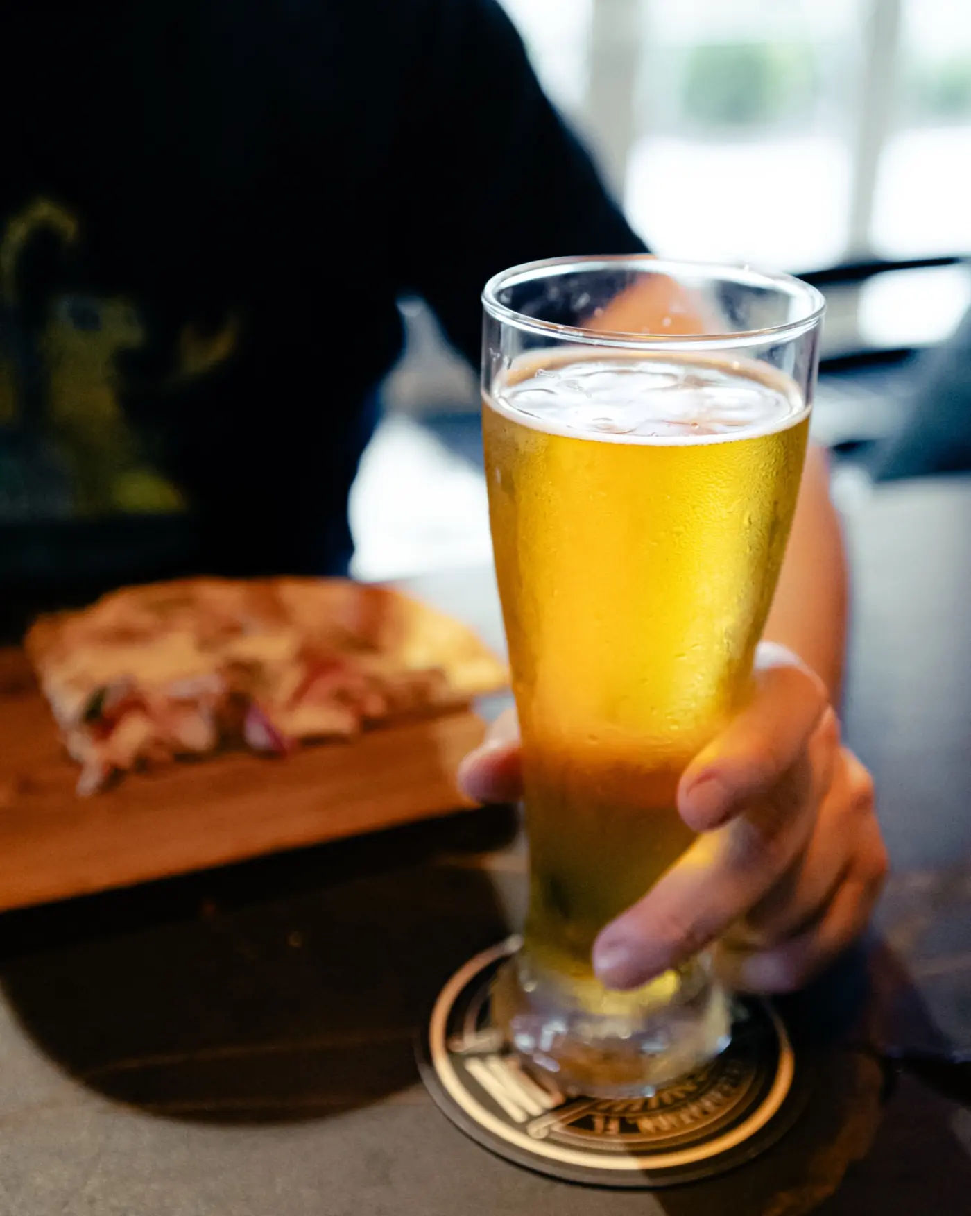 Customer holding a chilled pint of pale ale on a Celebration Brewing coaster, with a flatbread on a wooden board nearby